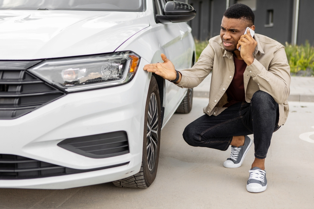A young black man sits near a car with a broken glass taking an appointment on the phone